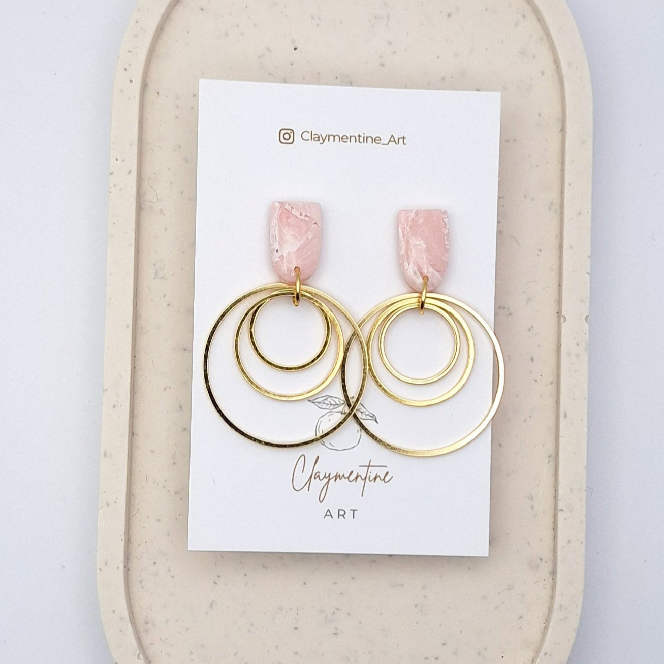 Gold hoop earrings with pink accents on a Claymentine Art card, displayed on a beige oval stand against a light gray background.